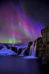 Colourful aurora across a star-filled night sky above waterfalls Water cascades over several rocky ledges
