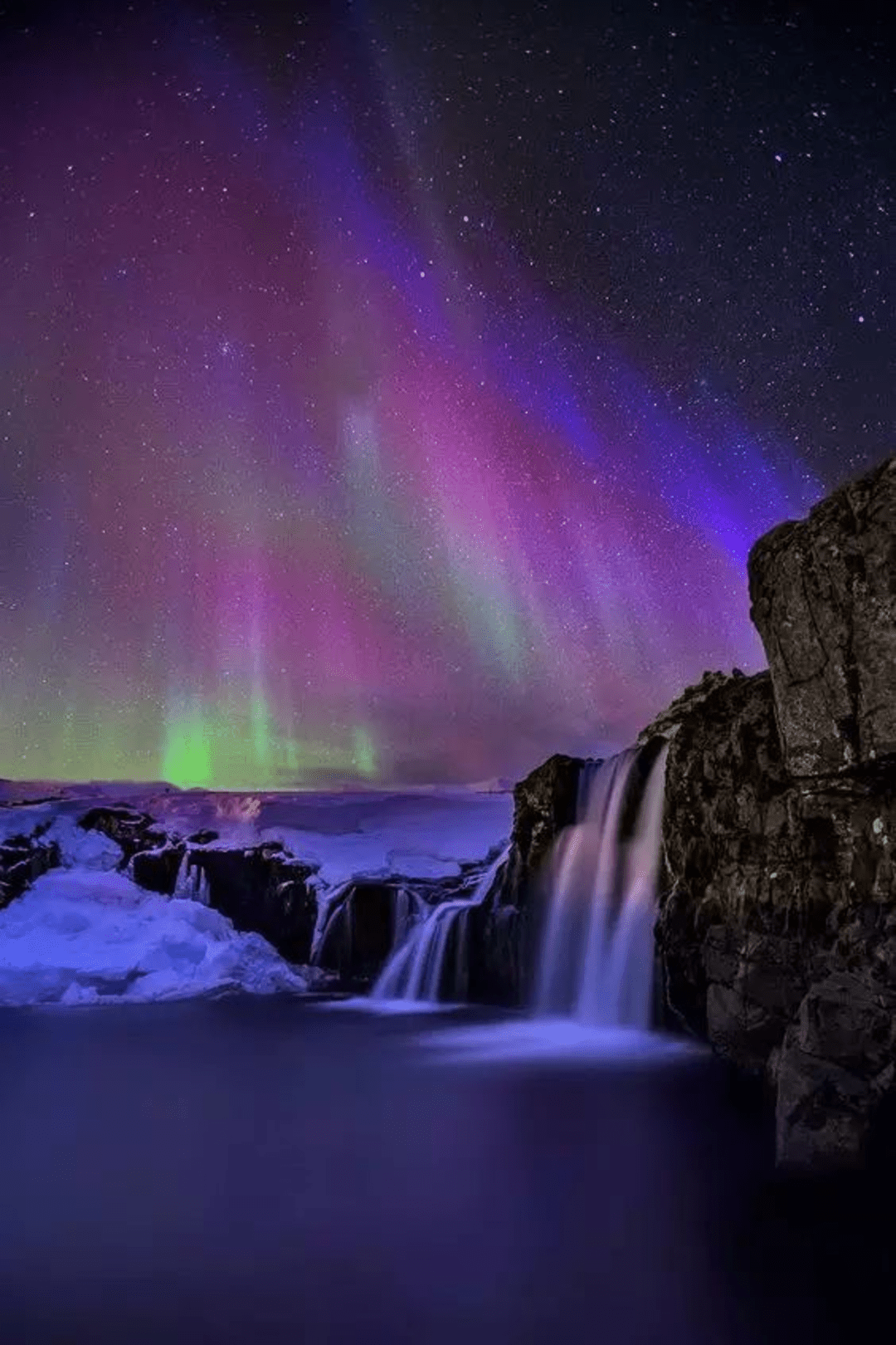 Colourful aurora across a star-filled night sky above waterfalls Water cascades over several rocky ledges